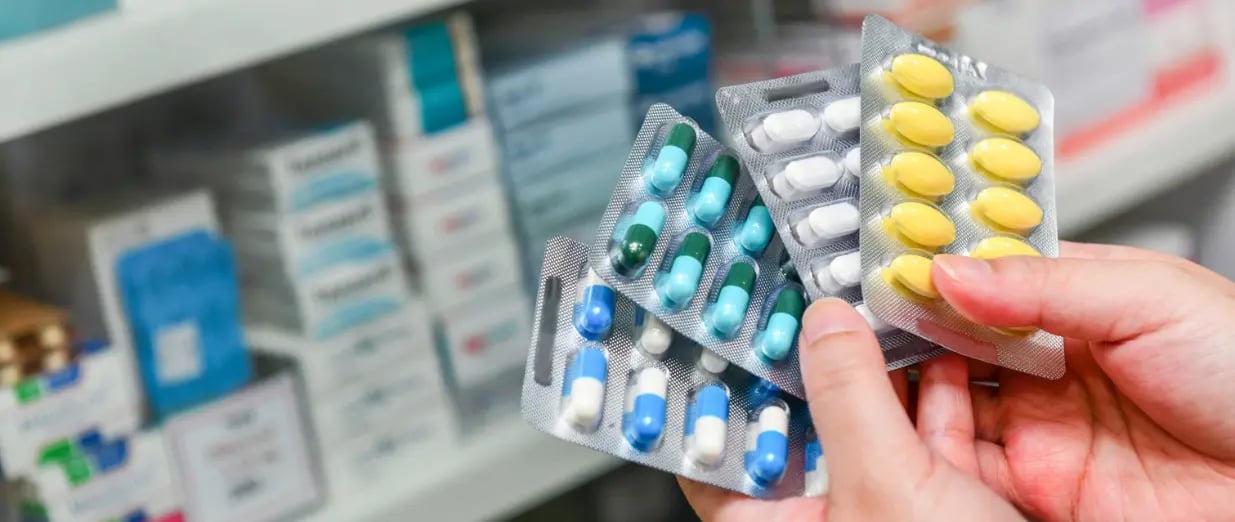 Hand h&auml;lt verschiedene Medikamenten-Blisterpackungen vor Apothekenregal