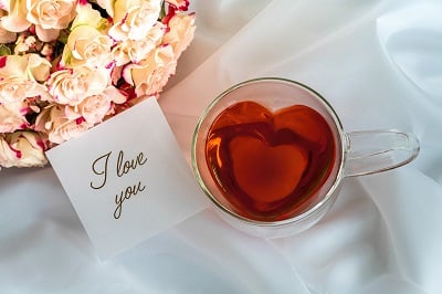 Teetasse aus Glas in Herzform, gefüllt mit Tee, rosa Rosen und einer Notiz mit den Worten "Ich liebe dich"