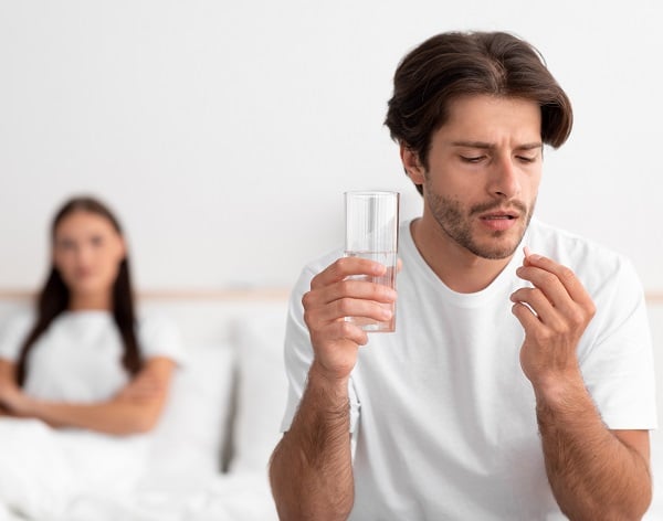 Ein Mann nimmt eine hellgelbe Pille Avanafil, während seine Frau im Bett wartet